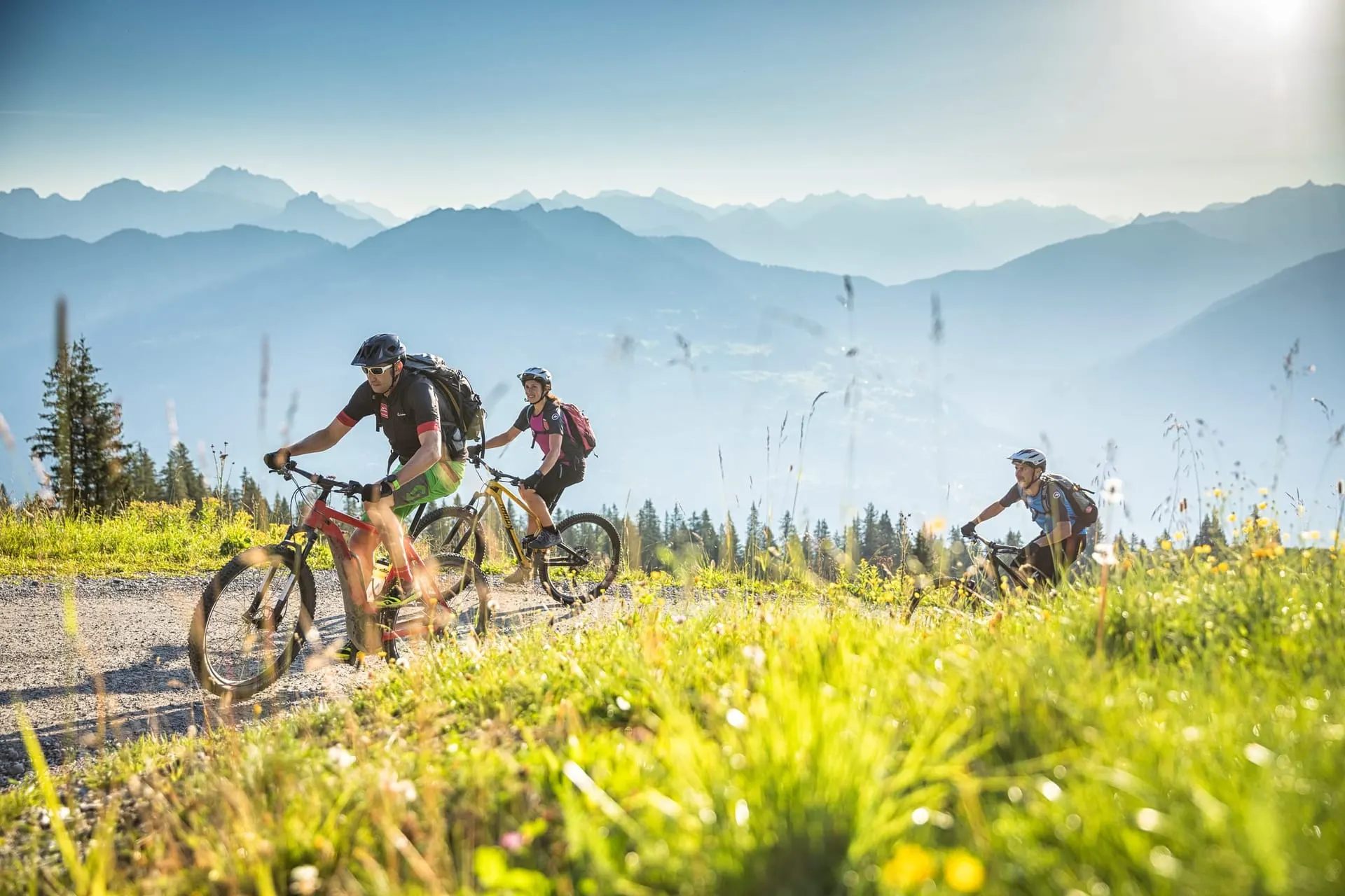 Three e-mountain bikers on a sunny mountain trail in the Montafon with a wide Alpine panorama in the background