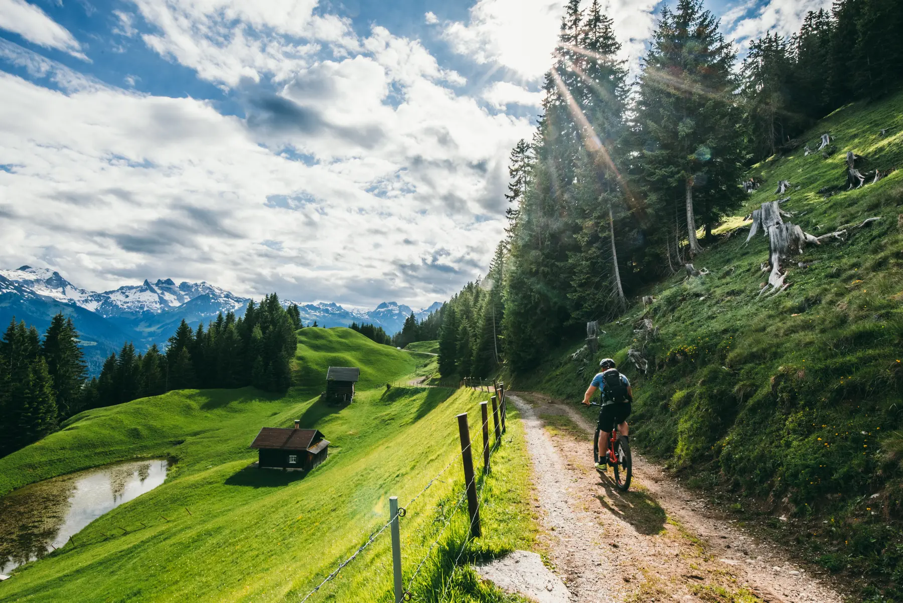 E-Mountainbike-Fahrer auf einem sonnigen Bergtrail rund um den Itonskopf im Montafon — Almwiesen und schneebedeckte Gipfel im Hintergrund
